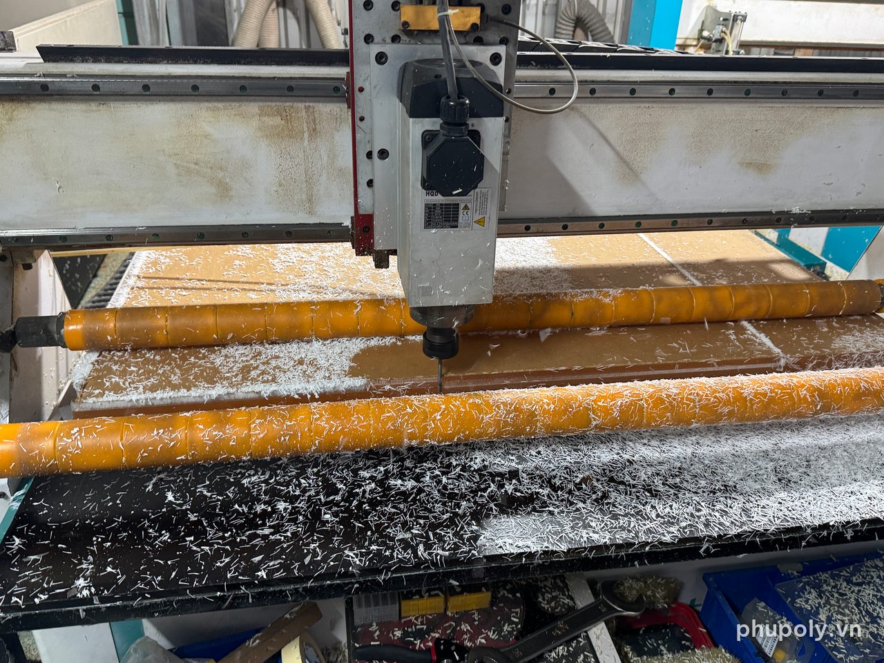 Đầu dao CNC đang cắt tấm mica 50mm còn giấy bảo vệ, phoi mica trắng phủ bàn máy