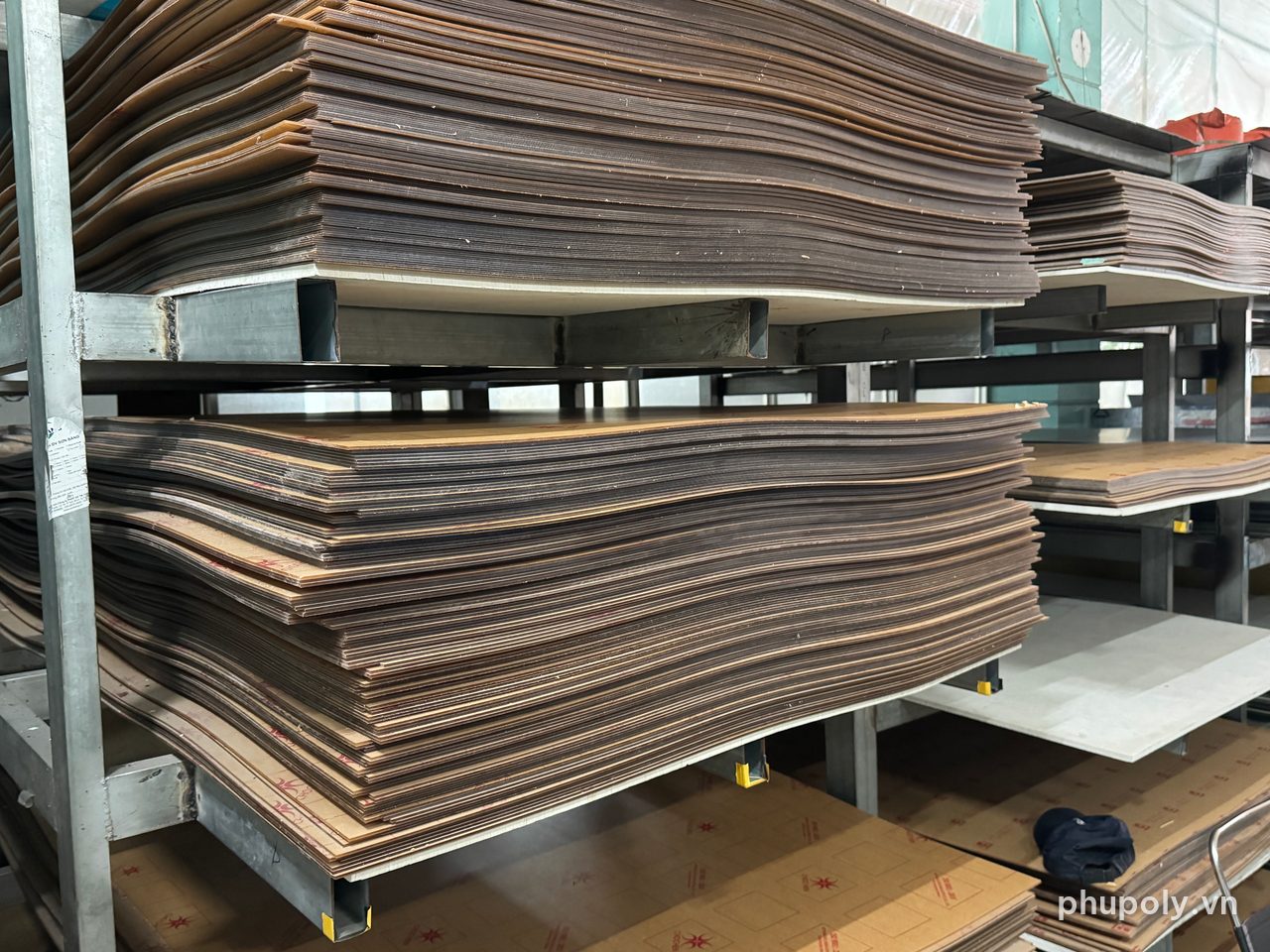 Chồng tấm mica còn giấy bảo vệ xếp nằm phẳng trên kệ sắt tại kho Phú Poly