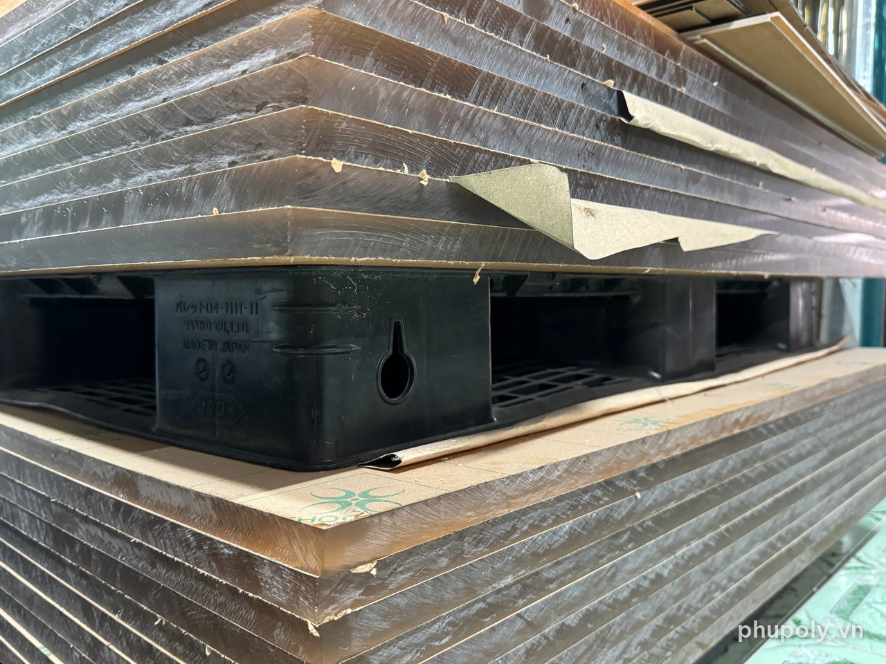 Cạnh các tấm mica dày còn giấy bảo vệ xếp nằm chồng trên pallet nhựa tại kho
